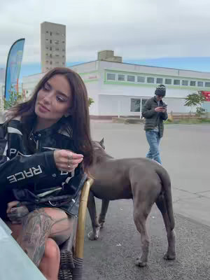A woman is sitting outside with a large grey dog, an older man is walking in the background. The woman is wearing a black jacket with a white logo, and has tattoos on her leg. The dog is standing and looking at her while she touches it gently.