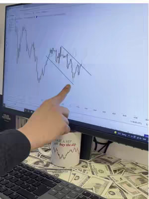 A person's hand points to a stock chart on a computer screen, with a keyboard and a mug that says 'Take a sip and buy the dip' in the foreground. Money is scattered across the desk.
