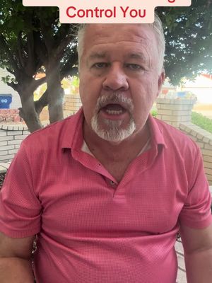 A middle-aged man with gray hair and a beard is speaking directly to the camera. He is wearing a pink polo shirt, and the background includes outdoor elements like trees, a brick wall, and a blue trash bin.