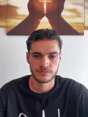 A man is looking directly at the camera. Behind him is a painting of two silhouetted hands holding a cross shape in a bright setting. The man has short dark hair, a beard, and is wearing a black shirt.