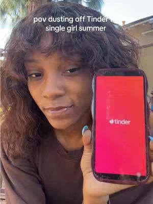 A young Black woman with wavy brown hair holds up a smartphone displaying the Tinder app. She's in front of an out-of-focus outdoor background.