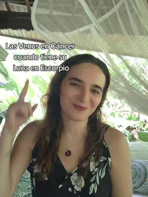 A woman with long, dark hair is pointing upwards with one finger. She is wearing a black and white floral top and a pendant. Text is overlaid at the top-left in white font on a dark background. The background shows greenery, appearing to be an outdoor setting.