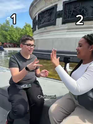The image shows two people outside with architectural stonework in the background. A person on the left is wearing glasses and a dark shirt, gesturing with their hands. On the right, a person is wearing a white shirt and holding a microphone, with one hand raised as if to give a high five. Text overlays read "1", "2", and "Chifoumi".