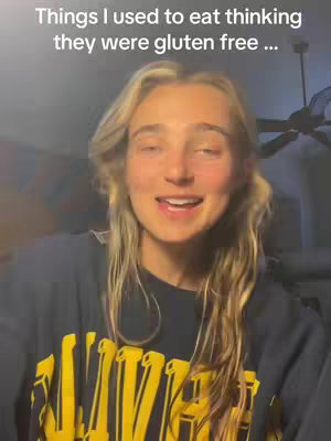 A woman with long blonde hair is smiling and talking to the camera. She is wearing a dark blue top with yellow lettering. Above her head is text that says, "Things I used to eat thinking they were gluten free..."