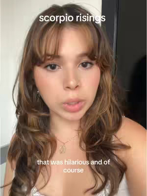 A woman with long brown hair and bangs is centered in the frame. Text overlays appear above and below, with text on the top reading 'scorpio risings' and the text on the bottom reading 'that was hilarious and of course'. She is wearing a small necklace and a white top. The image appears to be a selfie, shot from the chest up, with a neutral background.
