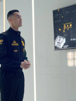 A man in a black jumpsuit stands in a brightly lit, minimalist room, looking towards the left. To his right, a screen displays technical diagrams and equations. The man is the focal point, with the background clean and uncluttered, creating a modern and professional aesthetic. The image has a futuristic, tech-oriented feel, enhanced by the neutral color palette and the geometric patterns on the wall.