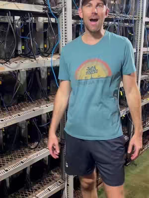 A man stands in front of racks of Bitcoin mining equipment. He is wearing a teal t-shirt, black shorts, and a baseball cap. He's speaking with an open mouth.