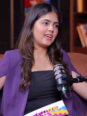 A young woman with long, brown hair sits in front of a microphone, likely in a podcast setting. She wears a black top and a purple blazer. The background is blurred, featuring a bookshelf and warm-toned accents, suggesting a home or studio environment.