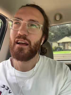 A man with a beard and glasses is speaking in the front seat of a car, with a suburban neighborhood in the background. The lighting is bright and cheerful.