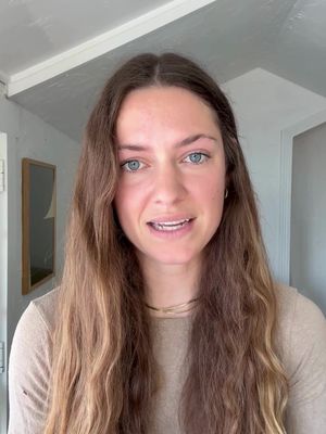 A woman with long, wavy brown hair and fair skin is centered in the frame. She is wearing a light beige top and a delicate gold necklace. Her eyes are blue and she is looking directly at the camera with a warm, open expression. The background includes a white ceiling and a doorframe with a framed picture, suggesting a casual indoor setting.