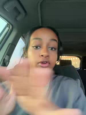 A person with dark hair and skin is filming from inside a car. The person is looking at the camera. The person's left hand is raised with fingers outstretched as if waving or gesturing. A blurry hand is visible near the face. The background is blurred, but parts of the car interior can be seen.