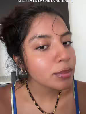 A close-up shot of a young woman's face, with a slight upward angle. Her dark hair is styled back. She's wearing a blue and white tank top and a necklace with a patterned pendant. Her skin appears lightly moist or dewy, with subtle makeup. She has a small nose piercing. The background is plain, and the lighting is natural. The focus is clearly on her face and expression.