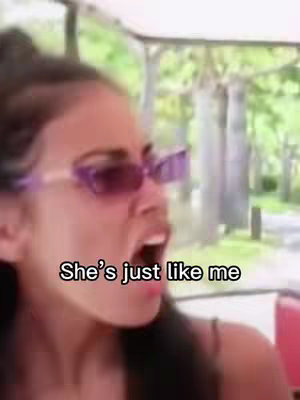 A selfie-style video cover image of medium shot of a woman with dark hair wearing purple-tinted rectangular sunglasses, eyes wide and mouth open in an expression of surprise or excitement. She appears to be outdoors as suggested by the blurred green background. The text overlay is a black text box with white lettering, reading , 'She's just like me'.
