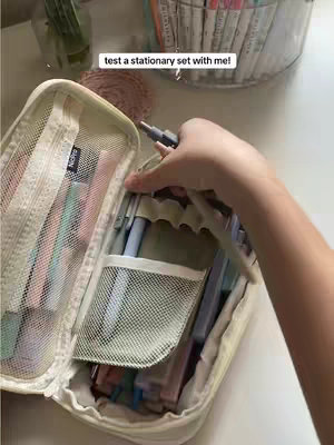 The image shows a close-up view of a person's hand opening a white, mesh-detailed pencil case. Inside the case are various colorful pens and markers. The background includes a desk surface, and a partially visible container of pens.