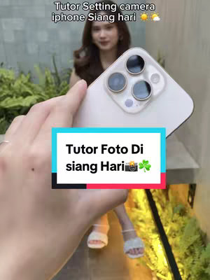 The cover image shows a person holding a white iPhone towards the camera. The phone's camera lenses are visible. A layered text box at the bottom has a light blue background with the text 'Tutor Foto Di siang Hari' on it, and a camera and clovers and other elements are also present. The background appears to be a garden setting.