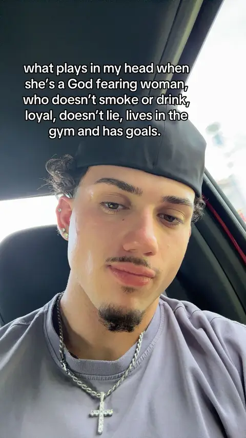 A young man with a slight smile and a goatee is seated in a car with a blurred background. He is wearing a gray t-shirt and a silver cross necklace. A black cap is on his head.
