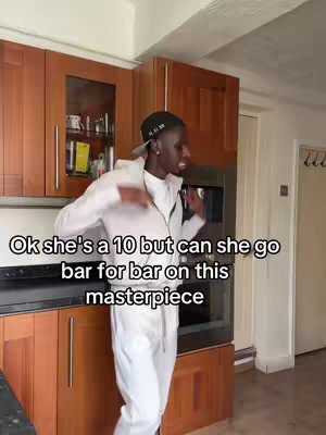 A man in a white tracksuit and a black cap is standing in a kitchen with wooden cabinets, gesturing as if he is dancing. Overlaid text in a white font on a black background says, 'Ok she's a 10 but can she go bar for bar on this masterpiece'.