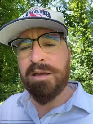 A close-up shot of a man wearing a baseball cap, glasses, and a beard. The background is a blurred view of green trees.