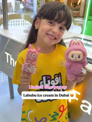 A young girl with dark hair smiles at the camera, holding a Labubu ice cream and a Labubu character doll. The girl wears a yellow t-shirt with Arabic text on it. The background is a store setting with displays.
