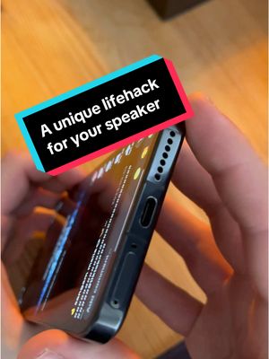 The cover image shows a close-up of a smartphone held in two hands. The phone's screen is visible, displaying an app interface. A rectangular text overlay in white font with a black background, is placed at the top of the phone with the text: "A unique lifehack for your speaker”. The image is set against a light brown wooden surface, with the phone angled slightly upward.