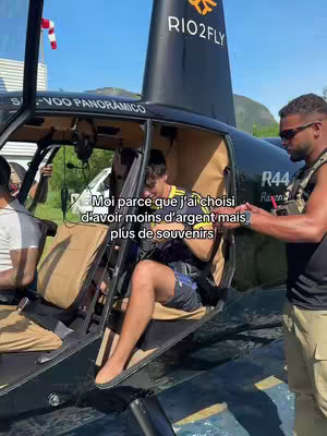 A man sits inside a black helicopter, with another man standing outside the open door. The man inside is wearing shorts and the other man is dressed in a vest. Text is overlaid in the image.