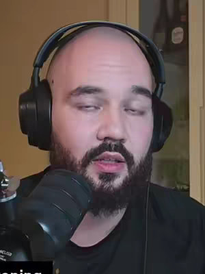 A man with a bald head, a full beard, and dark headphones is speaking into a microphone. The background is a light beige wall with a glimpse of a wooden door frame.
