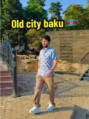 A full-length shot of a man standing on a paved area, with a blurred background featuring trees, a stone wall, and a stairway. The man is wearing a patterned shirt, brown pants, and white sneakers. Overlay text reads 'Old city baku' with an Azerbaijan flag icon.