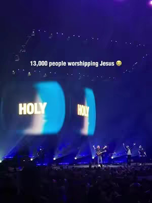 A concert stage with large screens displaying the word 'HOLY,' with a band performing in front of an audience, illuminated by a blue and purple light scheme.
