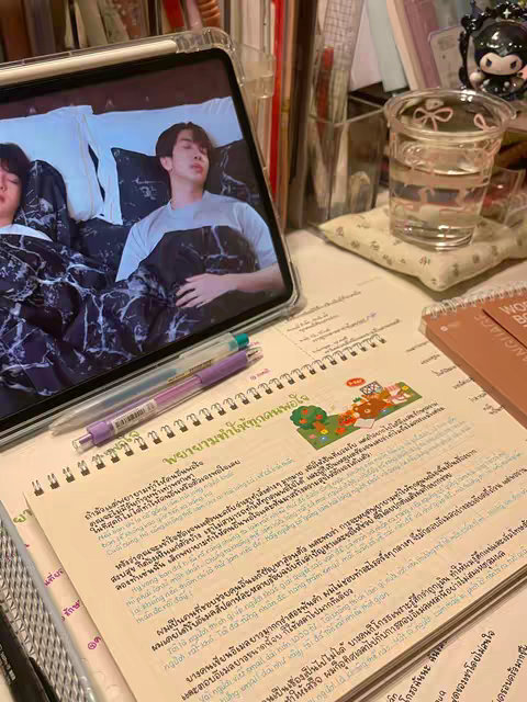 The cover image shows a workspace with a tablet displaying a scene of two men in bed, a notebook with handwritten text, and a clear glass of water. Pens are scattered on the desk, suggesting a study or work setup.