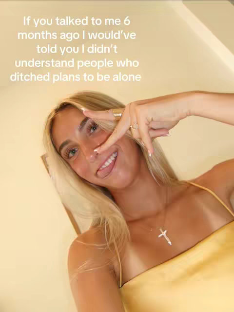 A woman with long blonde hair is shown from the chest up, looking at the camera with a smile. With her right hand she is making a peace sign near her eye. Text is overlaid on the image in a white font. The background is a peach color with some visible lighting.