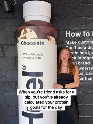 A woman stands next to a large prop of a Huel chocolate drink bottle. The woman has red hair and she is looking at the camera with a smirk. The background is a black wall with text on it.
