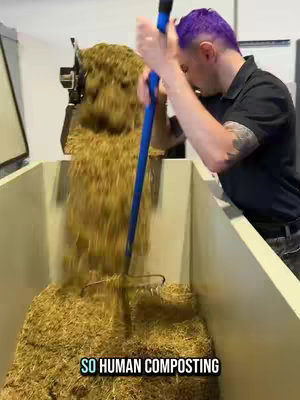 A man in black shirt with purple hair is using a rake to move brown matter into a large beige bin. The text overlay 'SO HUMAN COMPOSTING' appears in the lower portion of the video.