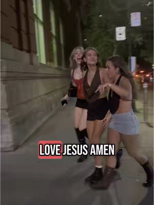 Three young women walking down a city street at night, with one pointing towards the camera. Text overlayed on the image in a red box reads 'LOVE JESUS AMEN'. The street has a dark, urban setting with a building and streetlights in the background.