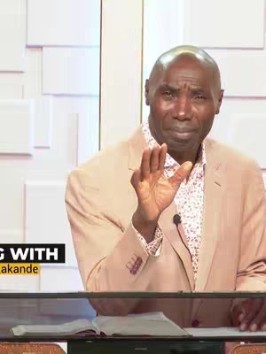 A man in a light beige suit jacket gestures with his right hand, palms up. The image has a soft color palette with light, neutral tones setting. The background appears to be a studio setting with simple, white walls.