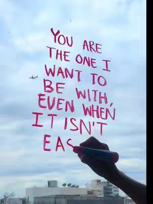 A hand holding a red marker writes the text 'YOU ARE THE ONE I WANT TO BE WITH, EVEN WHEN IT ISN'T EAS' on a clear window, with a cloudy sky and buildings visible in the background. An airplane is also visible in the sky.