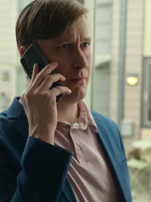 A man is talking on a phone looking concerned. He is wearing a blue blazer over a pink polo shirt. The background has a blurred view of an exterior setting.