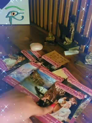 A dimly lit close-up shows a wooden surface scattered with tarot cards, a crystal, and decorative objects. A hand is reaching in to select or shuffle cards. The cards are in a spread, with some face up, revealing the colorful artwork specific to tarot cards. The top left has a painting of an eye, and the background features a wooden fence. The overall scene suggests fortune-telling activity.