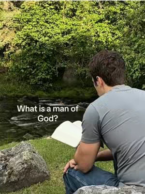 A man sitting by a stream, reading a book. The background is lush green foliage.