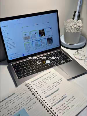 A well-lit workspace with a laptop displaying a white screen showing online learning documents, an open notebook with handwritten notes, and a decorative lamp on a white desk. The composition is clean, suggesting a study or work environment with focus on learning resources.