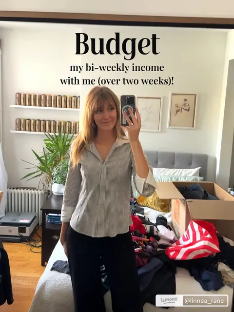 The cover image is a selfie taken in front of a mirror in a bedroom setting. The person, a young woman with light brown hair, is holding a phone in her hand. The text "Budget my bi-weekly income with me (over two weeks)!" is overlaid near the top of the image. The image has a slightly cluttered background with a bed, boxes, and various items visible.