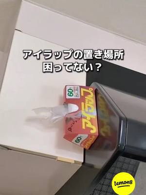 The image foreground features a wrap dispenser in open box form, clinging to a dark, curved surface that appears to be the side of a cabinet or an appliance. Above the dispenser, the text states, “アイラップの置き場所 困ってない?”, which translates to “Not having trouble finding a place for the i-wrap?”, the i-wrap is a Japanese brand to plastic wrap. The background includes a white cabinet or white wall.