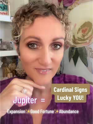 A woman with light skin and short curly hair is centered in a selfie-style shot. She gazes directly at the camera with a slight smile. Her left index finger points towards her jawline. She wears a purple top and a ring on her left ring finger. The background is slightly blurred but hints at a room with floral wall art.