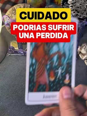 The image shows a hand holding a tarot card with a dark background. Overlayed on top is a yellow rectangle with the word "CUIDADO" in black text. Underneath, a red rectangle has the words "PODRÍAS SUFRIR UNA PERDIDA" in white text.
