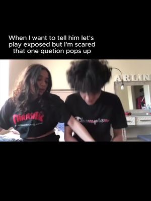 The image shows a medium shot of two young people, a woman and a man, in what seems to be an interior setting. The woman is on the left, with long, wavy, dark hair, wearing a black t-shirt with red text on it, and she appears to be looking down. Her head is angled towards the other person. The man is on the right, with curly dark hair, wearing a black Champion t-shirt, his head is down. The text is displayed with the upper half of the image. The setting is slightly blurred, suggesting they are in a home. Overall, the composition is somewhat intimate and likely related to a personal interaction.