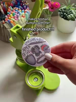 The cover image features a person's hand holding a round button maker with a finished product in a Christian theme. The button itself displays the "Armor of God" with illustrations and text referencing Ephesians 6:10-17. A green button-making machine is visible in the background, along with a container of colorful pens, a vase of flowers, and a succulent plant.