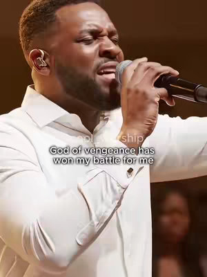 A man is singing into a microphone, dressed in a white dress shirt, with a text overlay and audience obscured by the low-resolution image.