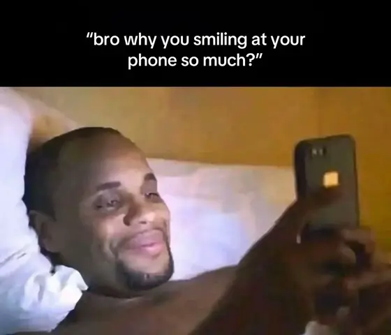 A man lies in bed, propped up on pillows, looking down at a phone that he is holding with both hands. He is smiling and appears to be engrossed in what he is viewing on the phone's screen. Overlaid at the top of the image is white text on a black banner.
