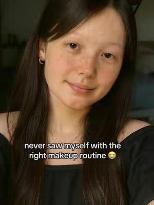 A selfie-style shot featuring a young woman with long, dark, wavy hair, light skin, and minimal makeup. She's positioned in the foreground with a softly blurred background, suggesting an indoor setting. The woman is smiling slightly and looking towards the camera. Text overlayed on the image reads, "never saw myself with the right makeup routine".