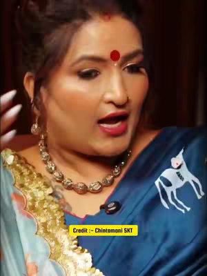 A medium shot of a woman with a red dot on her forehead, wearing a dark shiny top and a gold and silver necklace. She appears to be talking, with her mouth open. She is in front of a dark background. The text overlay is written with a yellow background.