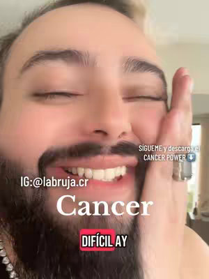 A selfie-style image with a man smiling widely, his eyes are closed in joy, and he is touching his cheek with one hand. Text overlays include the word 'Cancer' in a large, white font, and 'DIFÍCIL AY' in a red box underneath. At the top right, the text reads 'SIGUEME y descarga el CANCER POWER' with a blue arrow pointing downwards. The man has a beard, and the background appears to be an indoor setting.